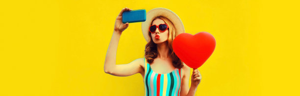 夏季肖像美丽的年轻女子自拍与智能手机与红色的心形状的气球戴墨镜在黄色的背景，空白的拷贝空间的广告文本图片下载