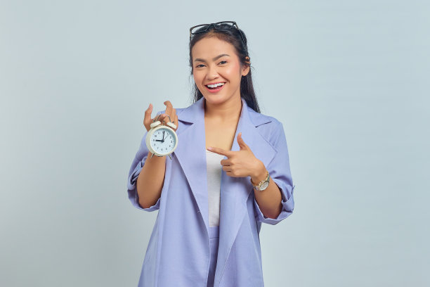 肖像开朗的年轻亚洲妇女指着闹钟孤立在白色背景图片下载