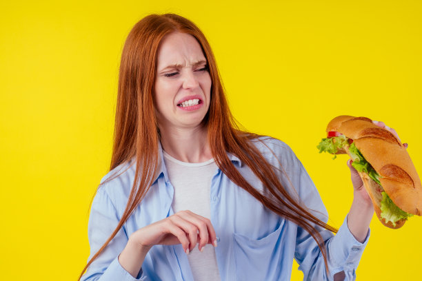 在工作室黄色的背景中，红发姜女拿着一个巨大的夹着胡椒和生菜沙拉的三明治图片下载