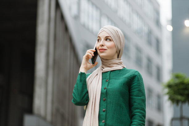 穿着绿色夹克和头巾的穆斯林女商人站在街上用手机聊天。图片下载