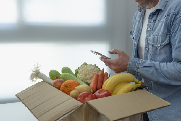 一名男子拿着智能手机在家里接收水果和蔬菜包裹。生态产品网购订单。纯素和素食的概念。图片下载