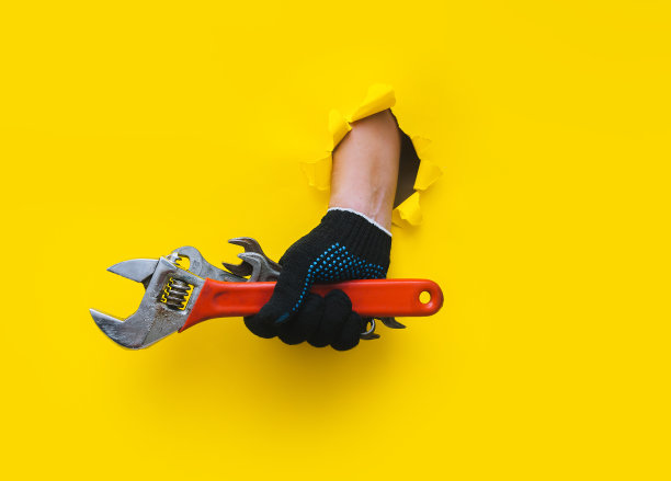 一只戴着黑色针织手套的右手，拿着几个扳手和一个红色的活动扳手。用黄色的纸撕破了一个洞。一个工人的概念，一个民工的概念，一个手艺大师的概念。副本的空间。图片下载