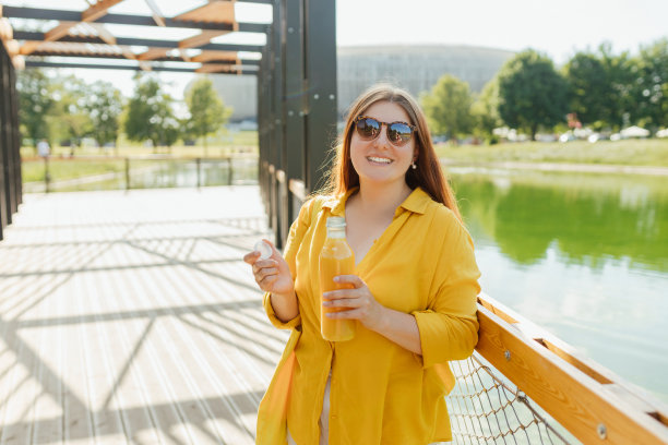 戴着墨镜的漂亮年轻女子在户外喝新鲜果汁。红发女孩走在公园里，拿着一瓶新鲜的冰沙在一个温暖的阳光明媚的日子。多维生素果汁瓶模型图片下载