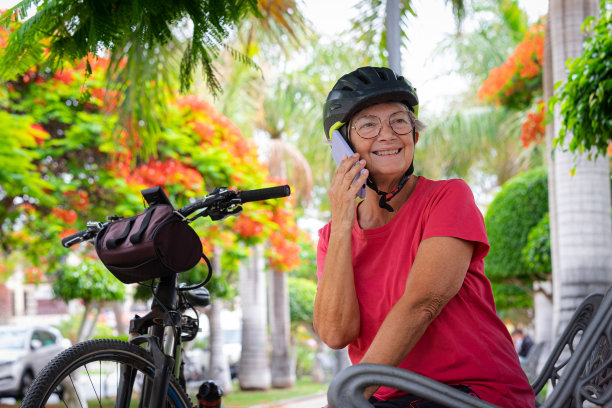 快乐的骑自行车的老妇人坐在公共公园的长椅上，靠近她的自行车，对着她的手机说话图片下载