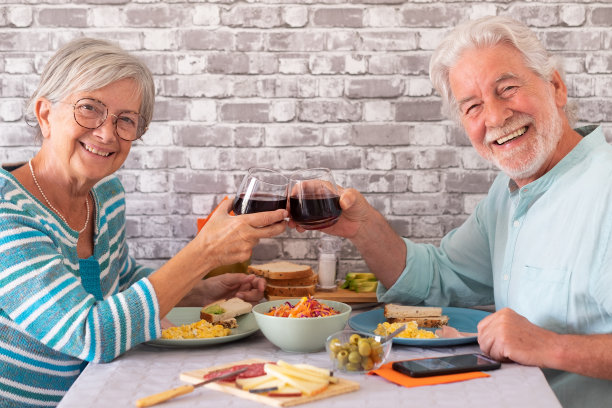 微笑的白人老年夫妇在家里面对面坐在桌子上吃早午餐时，用红酒杯敬酒图片下载