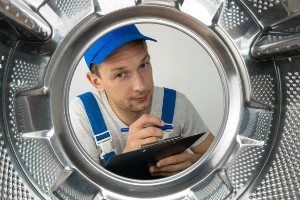 身穿制服的男修理工手里拿着一块平板，正在看洗衣机的滚筒，照片来自里面。图片下载