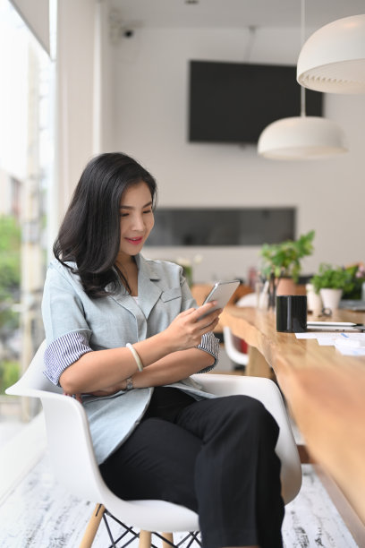 迷人的年轻女性坐在现代办公室，用智能手机在社交网络上聊天。图片下载