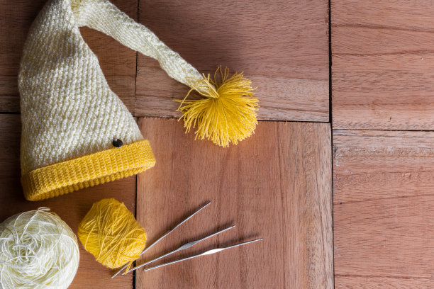 静物的黄色和白色的小精灵帽子为冬天，用钩针手工编织在一个木制的底色橙色上，旁边有两卷毛线和三根钢针准备在家里工作。图片下载