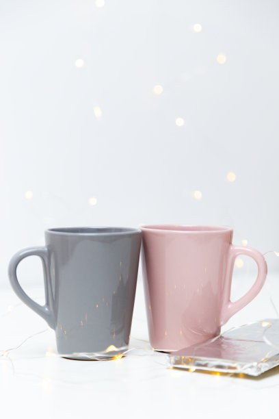 两个粉红色和灰色的咖啡杯模型和一块巧克力在白色的背景与小的金色灯光和bokeh。空白模板为您的设计，品牌，业务图片下载