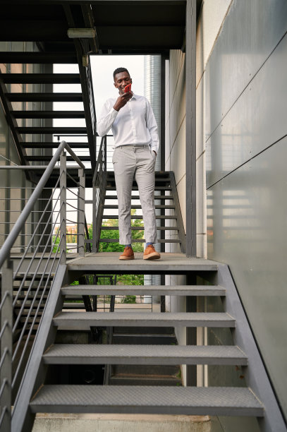 站在现代写字楼的楼梯上用智能手机发送语音信息的积极的非洲男性企业家的全身图片下载