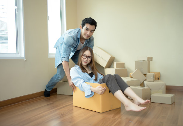 快乐的夫妇有乐趣，年轻的男人推年轻的女人坐在纸板箱看着镜头，愉快的室友玩，同时打包打开行李一起图片下载