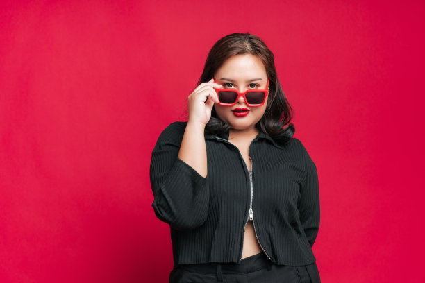 肖像美丽自信的亚洲女性加尺码戴着太阳镜在孤立的工作室红色背景黑色服装时尚摆拍。图片下载