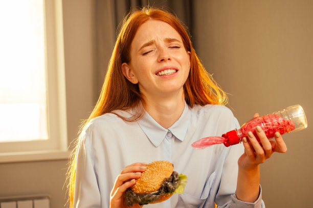 红发姜姑娘在家里的厨房里吃着包着芝麻的假汉堡和塑料薄膜垃圾，喝着用不环保管的一次性杯子里的水图片下载