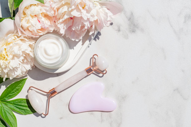 用于家庭护肤和水疗的面部套装。玫瑰石英面辊，刮沙按摩器，和一罐面霜在白色大理石背景牡丹花。自然治疗的概念。前视图。图片下载