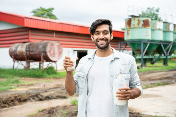 白人男性奶农在牛棚中手持牛奶瓶的肖像。年轻英俊的农业男性农民微笑着看着镜头后，在畜牧业幸福挤奶图片下载