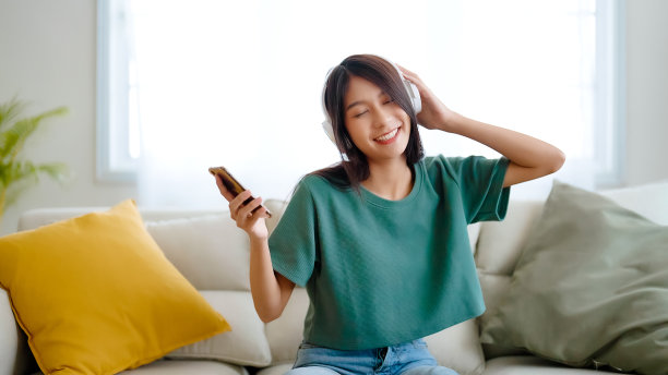 年轻的亚洲女性在家里客厅的沙发上听音乐。快乐亚洲女性使用手机，戴耳机，坐在沙发上图片下载