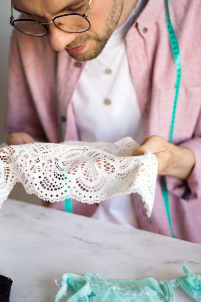 一个穿着粉色衬衫的裁缝的特写。一位戴眼镜的男设计师为内衣选择蕾丝的类型。一个脖子上挂着卷尺自学成才的裁缝。生活方式的概念图片下载