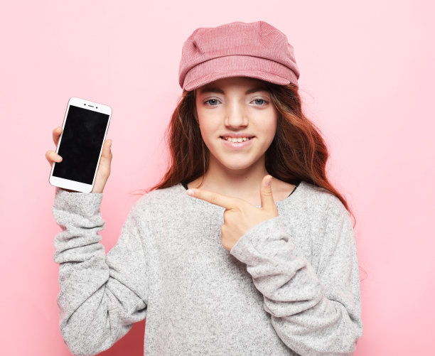 特写肖像开朗的女孩戴着粉红色的帽子显示智能手机孤立在粉红色的背景图片下载