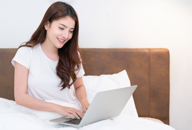 快乐休闲的漂亮女人坐在房子的床上，在笔记本电脑上工作。快乐的女人读新闻，她在床上放松与笔记本电脑在早上图片下载