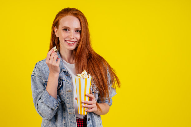 情绪激动的红发姜女穿着牛仔裤夹克，在工作室黄色的背景中拿着爆米花图片下载