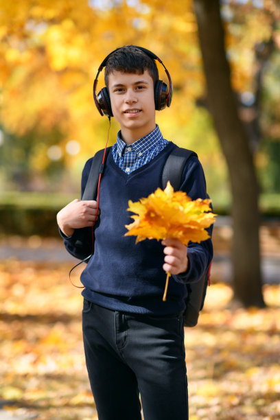 一个十几岁的男孩走在秋天的公园里，戴着耳机听着音乐，阳光明媚的日子里，树上的叶子都泛黄了图片下载