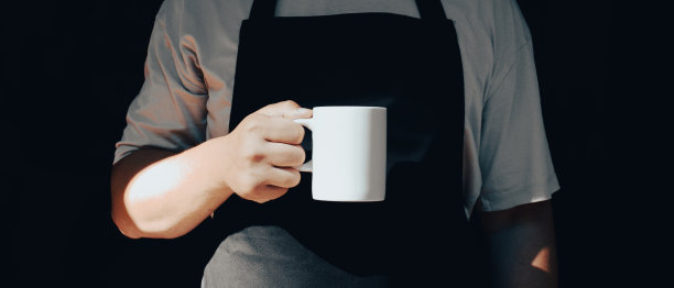 在黑暗的背景下，穿着围裙的男人手里拿着一杯咖啡。咖啡店横幅设计。图片下载