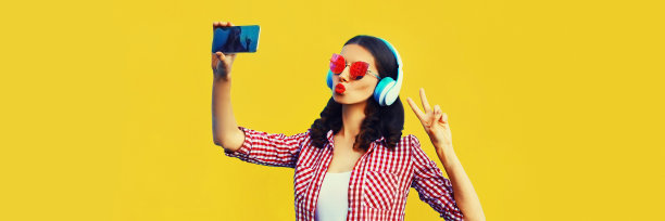 时尚年轻女性的肖像自拍与智能手机听音乐耳机和吹她的嘴唇发送甜蜜的空气吻在黄色的背景图片下载