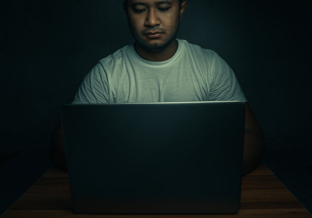 在一间黑暗的房间里，一个穿着白衬衫的男人坐在一台笔记本电脑上。有光照耀着。互联网addiction.topview概念。图片下载