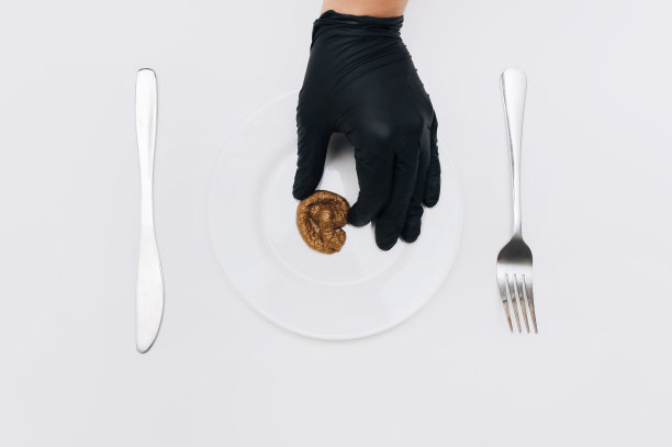一只戴着黑色手套的左手把假粪便放在盘子里，盘子上放着一张餐叉和一把刀。假新闻的概念，卑鄙，糟糕的食物，和糟糕的笑话。图片下载