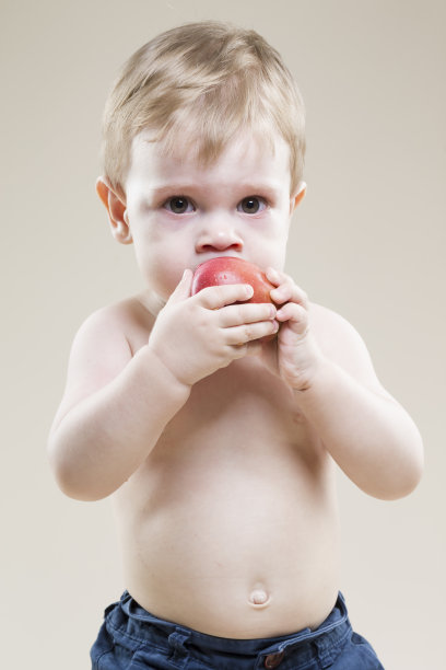 一个微笑的小孩子手里拿着食物的自然肖像，在米色背景下吃红苹果。图片下载
