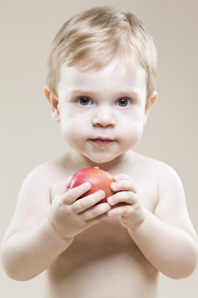 一个微笑的孩子手里拿着食物，贪婪地吃着红苹果，在米黄色的背景下摆姿势的自然肖像特写图片下载