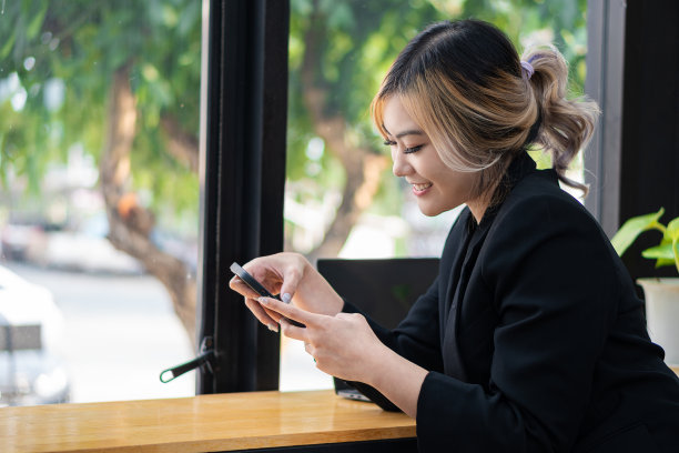 漂亮的亚洲女人坐在她的办公桌前微笑着讲手机和玩笔记本电脑。分享好的商业故事图片下载