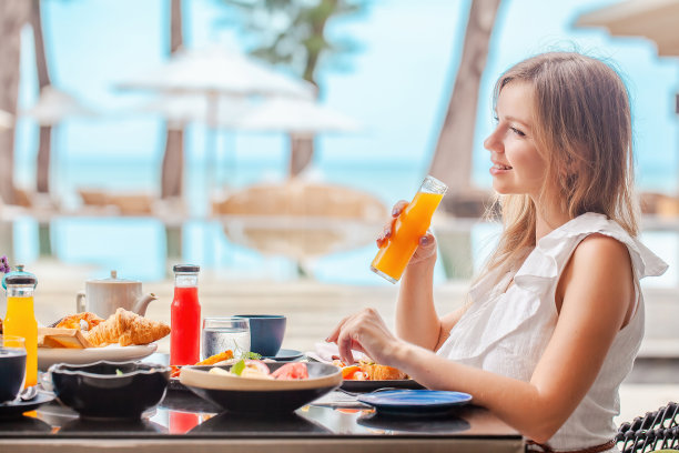 在豪华的热带度假村的游泳池附近喝健康橙汁的早餐妇女。泳池旁的健康早餐和水果盘。海滩奢华生活，夏日度假。图片下载