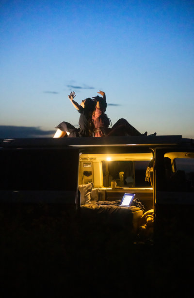 两个女人晚上坐在面包车的车顶上，背靠着背，双臂张开，面包车的车门被打开，车内灯火通明图片下载