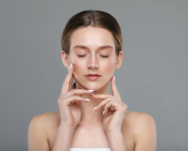 美丽的年轻女子干净新鲜的皮肤和闭上的眼睛触摸她的脸在灰色的背景。图片下载