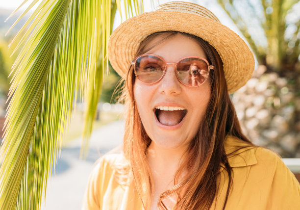 戴着草帽和墨镜的快乐红发女子在热带岛屿度假时自拍，背景是棕榈树。女性独自旅行。夏天的概念。图片下载