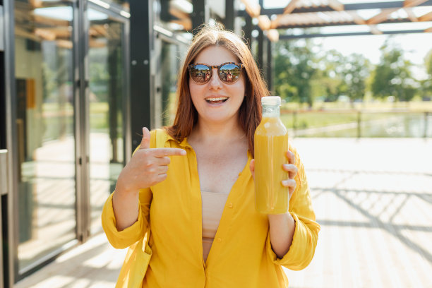戴着墨镜的漂亮年轻女子在户外喝新鲜果汁。红发女孩走在公园里，拿着一瓶新鲜的冰沙在一个温暖的阳光明媚的日子。多维生素果汁瓶模型图片下载