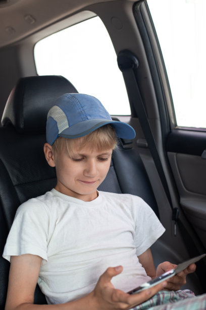 一个戴着帽子的男孩坐在汽车后座上，看着手机屏幕，微笑着图片下载