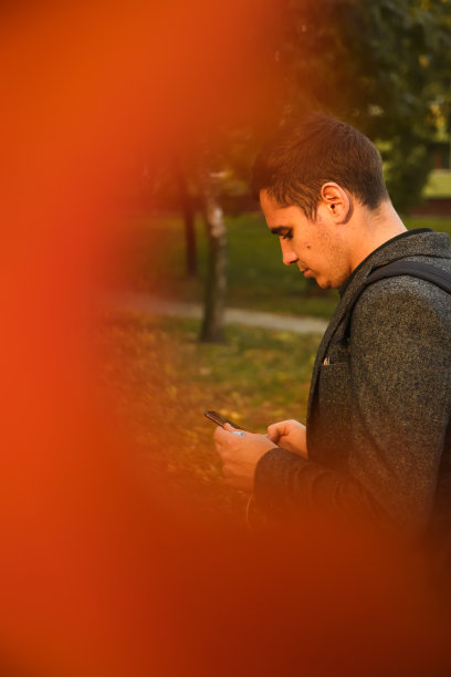 分散注意力的年轻人拿着电话。一名男子在秋天的公园里使用智能手机。用手机输入短信或阅读社交媒体。秋天的背景。手机。红色的框架。沟通。的焦点图片下载