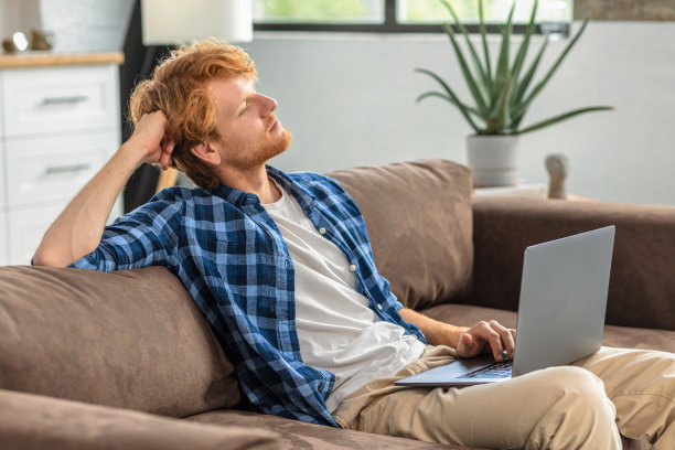 无忧无虑的年轻人坐在家里的沙发上，看着窗外，想着一些事情。年轻的白人商人或自由职业者，正在休息，放松图片下载