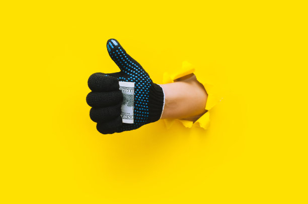 戴着黑色工作手套的右手拿着美钞(钱)，并竖起大拇指(像这样)。用黄色的纸撕破了一个洞。外来工、杂工、工资制的概念。副本的空间。图片下载