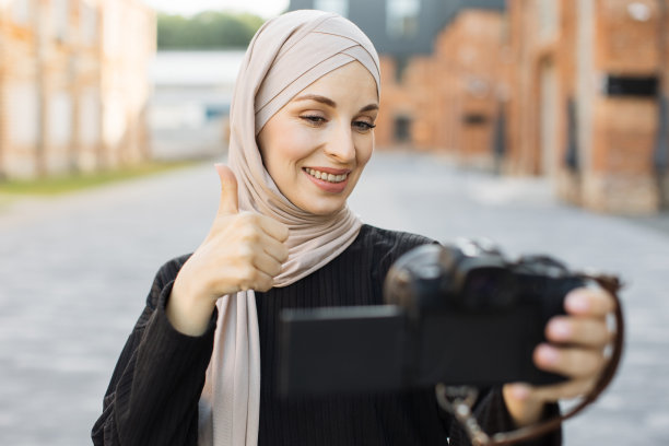 高兴和兴奋的阿拉伯女孩旅行者穿着黑色的衣服和希贾布，向互联网观众挥手。图片下载