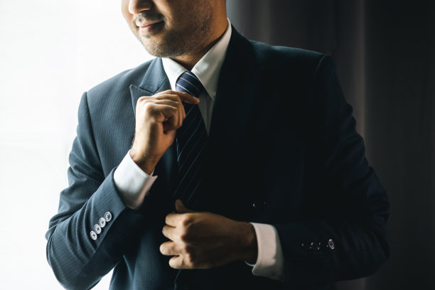 自信的商人在酒店房间的窗边用领带调整经典的蓝色西装。一个帅哥在婚礼上穿了一套漂亮的西装。图片下载