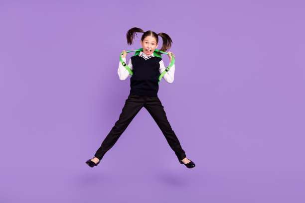 照片运动可爱的小学生女孩跳有乐趣穿背包统一孤立的紫色背景图片下载