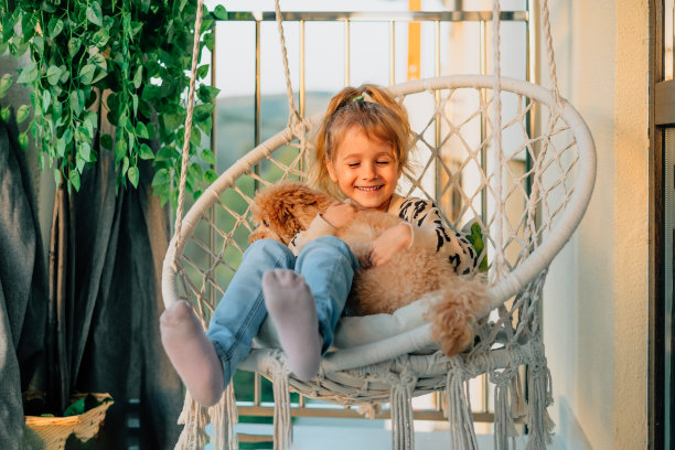 快乐的小女孩，孩子微笑着拥抱着她的宠物，狮子狗春天在家里的阳台上，夏天在棉流苏吊床的椅子上日落。图片下载