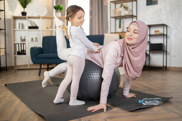 一位穆斯林妇女正在用健身球伸展身体，她迷人的小女儿站在垫子上图片下载