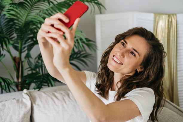 这个女孩手里拿着一个手机。一个女人在互联网上写信息，看图片，搜索信息。女孩为社交网络自拍。女人微笑着坐在家里的沙发上放松图片下载