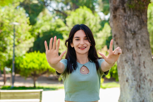 开朗的黑发女子穿着t恤站在城市公园，在户外用7号手指指着，微笑着自信而快乐。选择性聚焦在她的脸上。图片下载