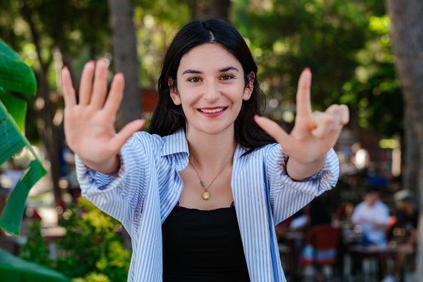 快乐的女人穿着条纹衬衫站在城市公园，户外显示和手指7，微笑着自信和快乐。选择性聚焦在她的脸上。图片下载