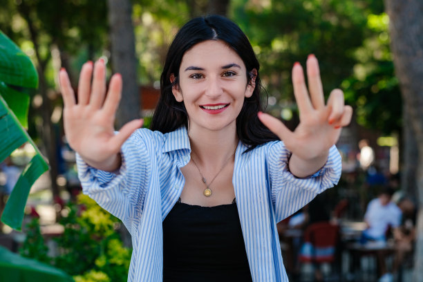 快乐的女人穿着条纹衬衫站在城市公园，户外显示和手指8，微笑着自信和快乐。选择性聚焦在她的脸上。图片下载